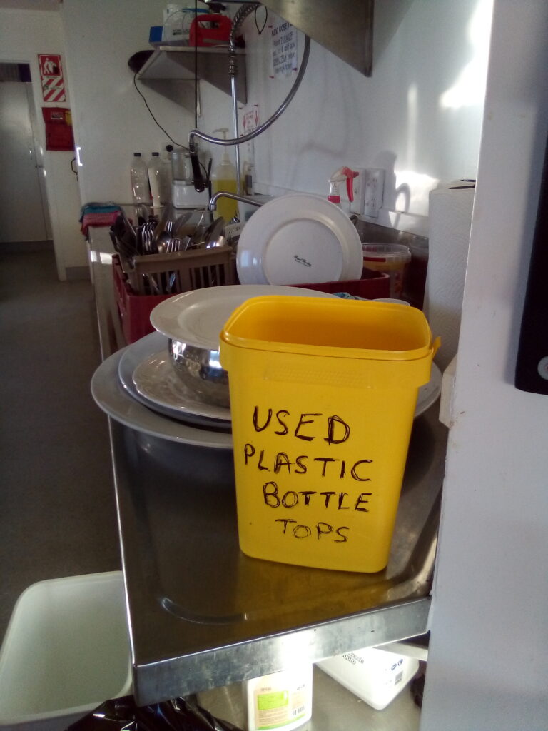 11. Ensure Proper Waste Bin Placement and Signage<br>11. Cardboard boxes housing beverages, food, and supplies were flattened and placed in a designated reclamation bin. (Photo 11a) Smaller WOWW-specific waste bins were created and placed in appropriately-marked baskets in more than one location for transfer to bigger bins for waste management pick up. (Photo 11b) Bottle caps were sorted in the kitchen and at the bar for club recycling or local school fundraising. (Photo 11c)  