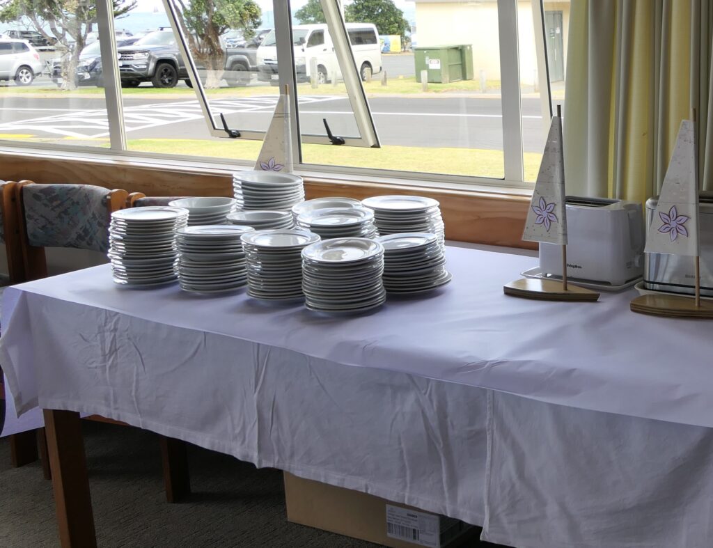 3. Serve Food with Plastic-Free Dinnerware<br>3. Gulf Harbour Yacht Club has always used plastic-free beverage glasses, china, and cutlery for its food and bar services.During the 2025 WOWW event, we reaffirmed the club’s commitment to plastic-free food and bar service with caterers and volunteers. Morning coffee service was supplied with china mugs, metal teaspoons, and china cream pitchers. (Photo 3a) China plates and bowls, and cloth tablecloths, were used for all food service. (Photo 3b) Lunches on participating boats were  provided in bags made of hessian with paper labels and cotton strings. Sandwiches were wrapped in wax paper and fresh fruit required no wrapping. Prizegiving Night tables were set with linen tablecloths, real glasses, metal cutlery and small paper napkins which were composted after use. Centerpieces were reused. (Photo 3c) Ends of roll paper from printing were repurposed on top of buffet tablecloths to reduce the number of times they needed to be laundered. (Photo 3d)